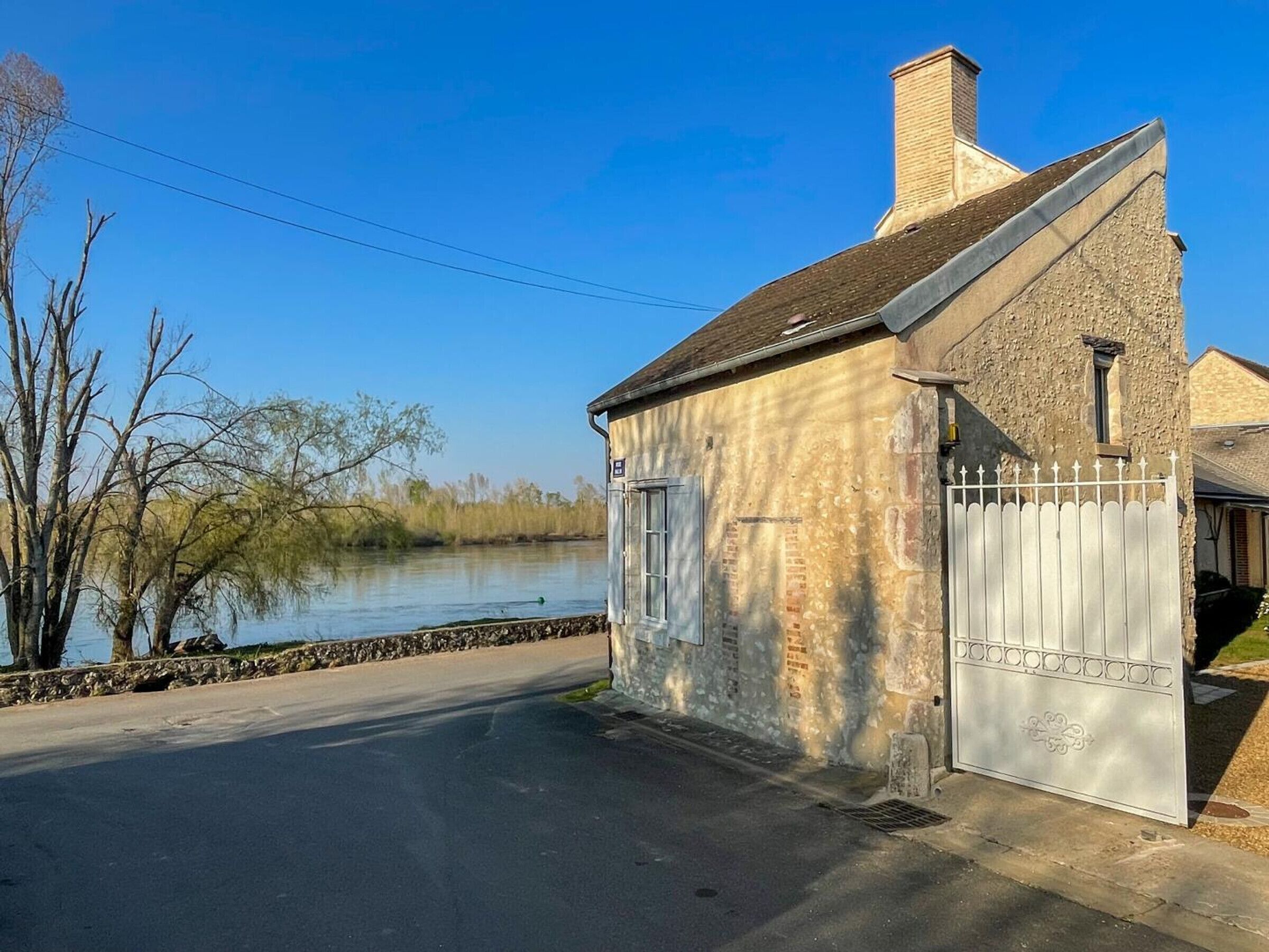 Un petit bâtiment en pierre avec une porte blanche, un ciel dégagé et une étendue d’eau en arrière-plan.