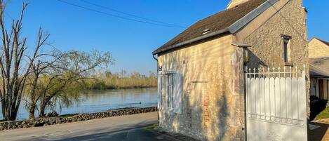 Un petit bâtiment en pierre avec une porte blanche, un ciel dégagé et une étendue d’eau en arrière-plan.