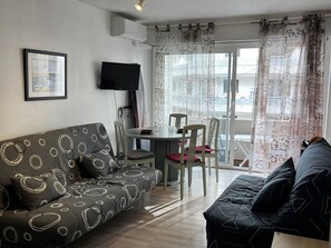 Ein Wohnzimmer mit Sofa, einem Esstisch mit Stühlen, einem Flachbildfernseher und einem Balkon mit gemusterten Vorhängen.