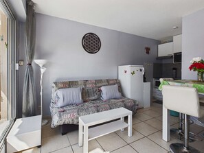 A compact living space with a sofa, a small table, a chair, a refrigerator, and a kitchen area. - Spacious apartment with parking - 4 guests (Marseillan-Plage)