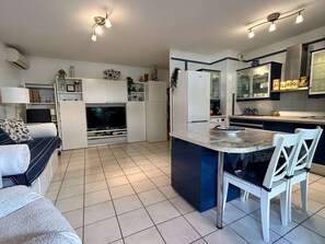A modern kitchen with a central island, built-in appliances, and a dining area with white chairs. - Modern apartment with parking and wifi (Balaruc-les-Bains)