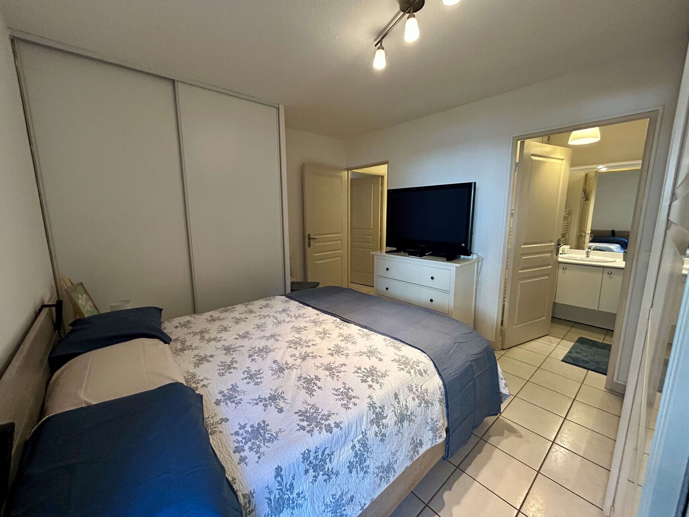Een slaapkamer met een bed, een televisie op een dressoir, een badkamer met een wastafel en spiegel, en een deur die naar een andere kamer leidt.