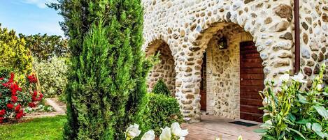 En byggnad av sten med välvda dörröppningar, omgiven av frodig grönska och blommande växter.