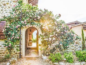 Un edificio de piedra con una entrada en arco, rodeado de abundante vegetación y plantas florecientes.