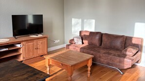 A living room with a flat-screen TV, wooden furniture, a brown leather sofa, and a window with natural light. - Guesthouse LeLo (5) (Solingen)