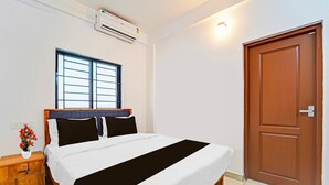 A bedroom with a bed, a window, an air conditioning unit, a wooden nightstand, and a door. - Deluxe Kalamassery Near Medical College (Kanayannur)