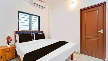 A bedroom with a bed, a window, an air conditioning unit, a wooden nightstand, and a door.