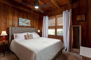 A wooden-paneled bedroom with a bed, two bedside lamps, a ceiling fan, and a window with curtains. - Ocean Views + Steps to the Beach + 3BDR + King Bed (Oak Island)