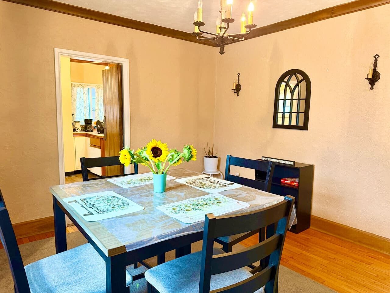 Een eetruimte met een tafel gedekt voor vier personen, een kroonluchter, een raam met decoratieve omlijsting en een deur naar een andere kamer.
