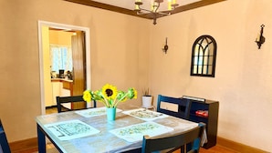 Un comedor con una mesa preparada para cuatro personas, una araña, una ventana con marco decorativo y una puerta que conduce a otra habitación.