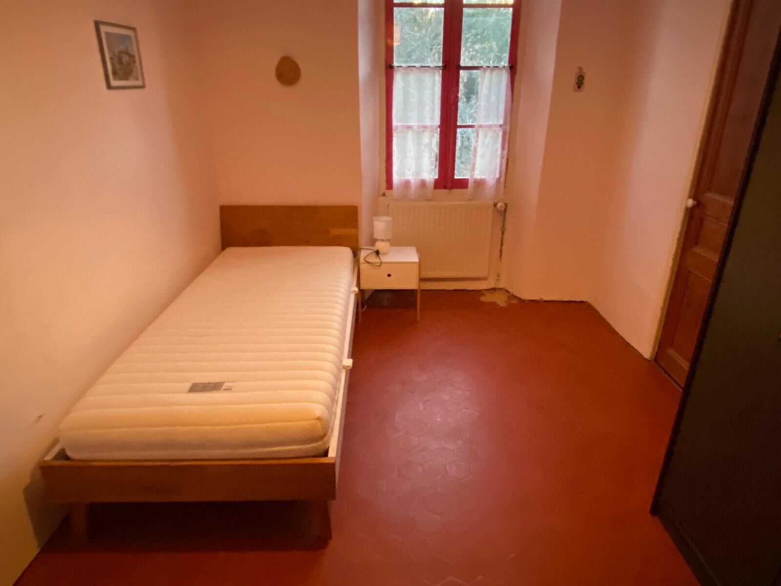 A small, single-bed room with a wooden headboard, a bedside table, a radiator, and a window with curtains.