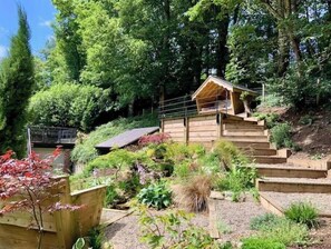 Eine Holzhütte mit geneigtem Dach, umgeben von einem Garten mit verschiedenen Pflanzen und einer kleinen Brücke.