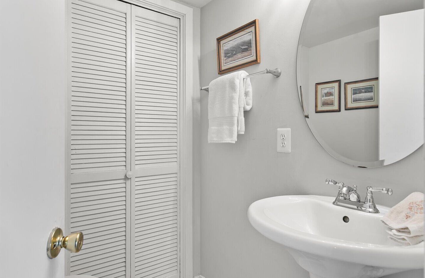 A bathroom with a white sink, a round mirror, and a white door with horizontal blinds.
