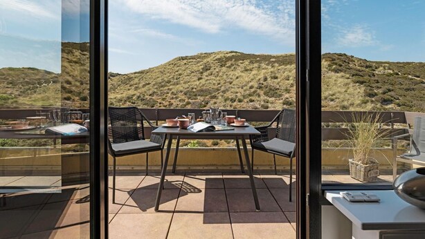 Una terraza con mesa y sillas, vista a un paisaje montañoso y un ventanal con el reflejo de la escena.