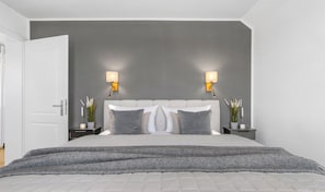 A bedroom with a bed, two bedside tables, a nightstand, a door, and a wall-mounted lamp. - Deichvilla Weitblick – Exclusive New-Build with Unobstructed Dike Views (Büsum)