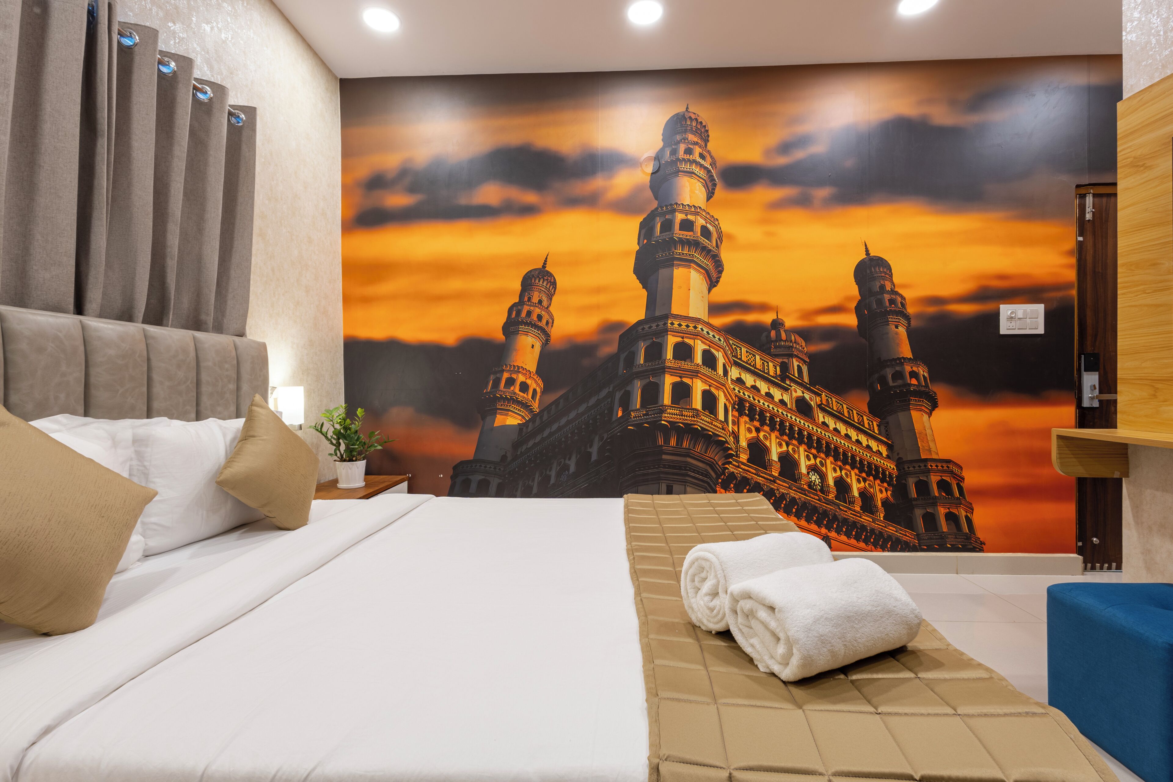 A hotel room with a bed, a wall-mounted TV, a towel on the bed, and a wall mural of a building.