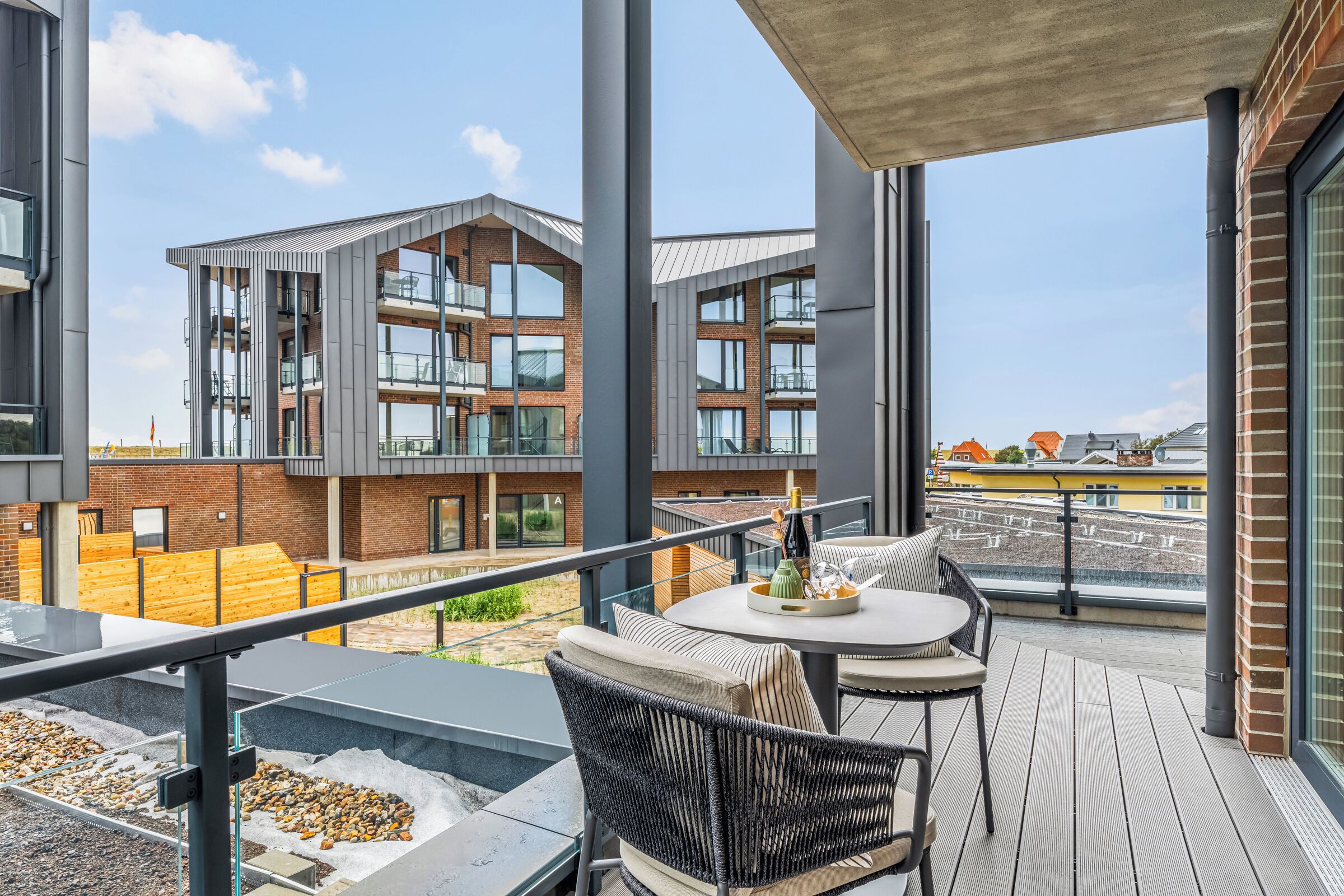 A modern outdoor balcony with a seating area, a view of adjacent buildings, and a clear sky.