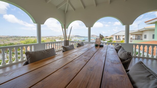 A wooden table with benches and cushions on a covered patio with white railings, overlooking a landscape with hills and a building in the distance. - The View The Ambassador (Jan Thiel)