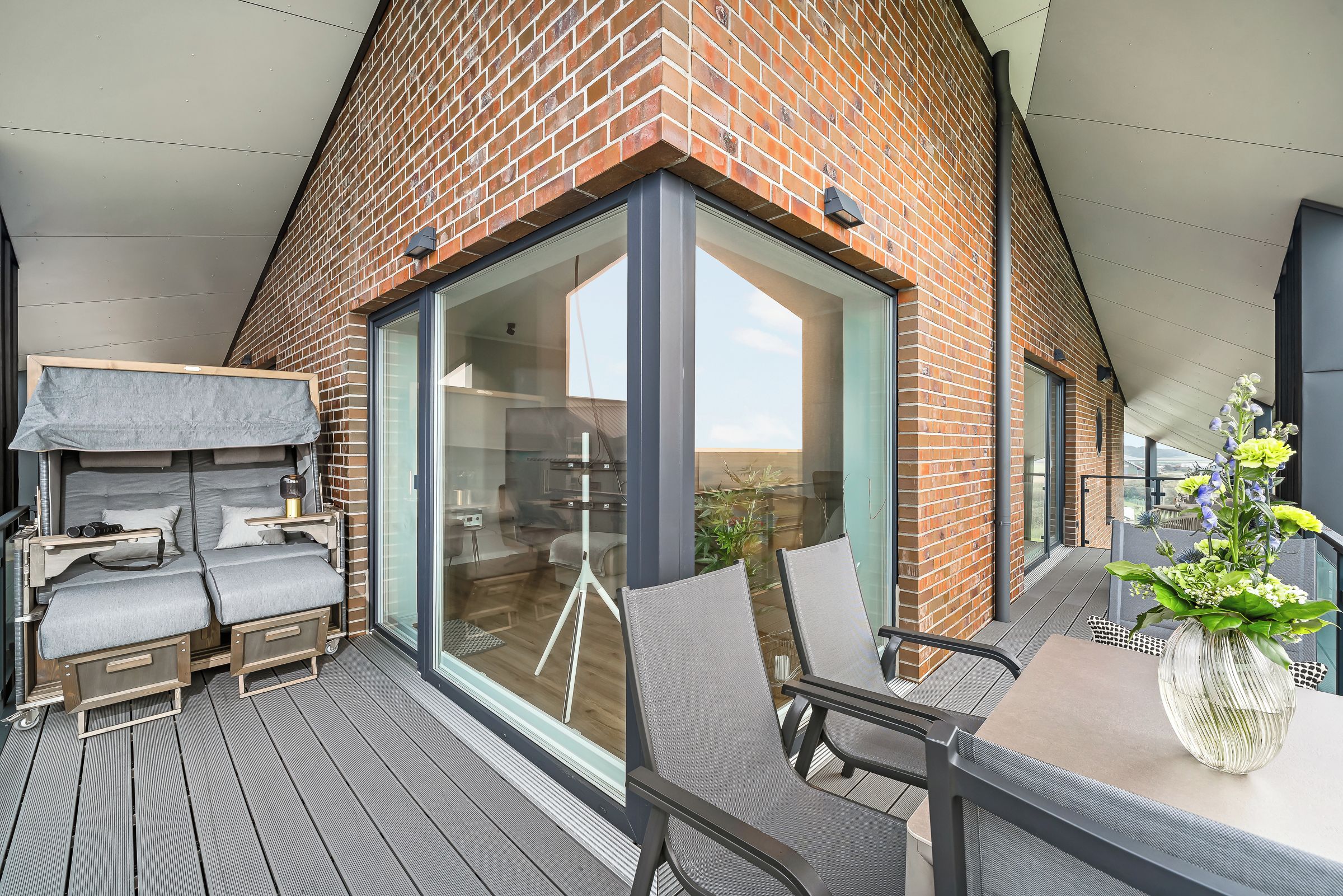 A modern outdoor balcony with a glass door leading to a room, furnished with a sofa, chairs, and a table.