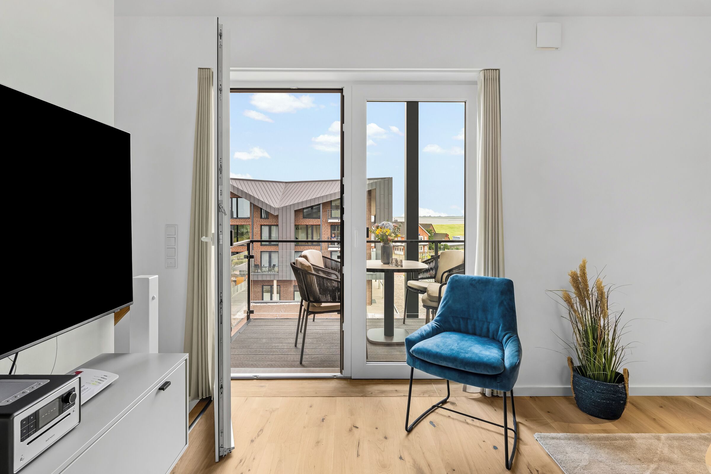 A modern living room with a television, a blue chair, a potted plant, and a sliding glass door leading to a balcony with outdoor furniture.