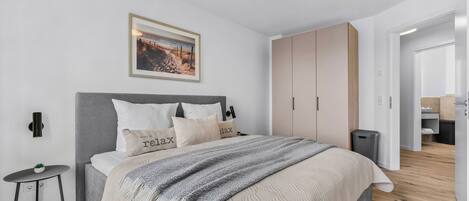 A modern bedroom with a bed, a bedside table, a wall-mounted lamp, a framed picture, and a wardrobe.