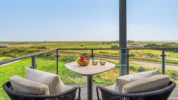 Een balkon met rieten meubels, een tafel met een schaal fruit en twee kommen, en uitzicht op een groen landschap met een spoorlijn.