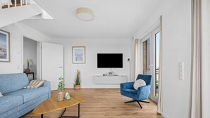 Een moderne woonkamer met een blauwe bank, een houten salontafel, een blauwe fauteuil en een flatscreen televisie die aan de muur bevestigd is.
