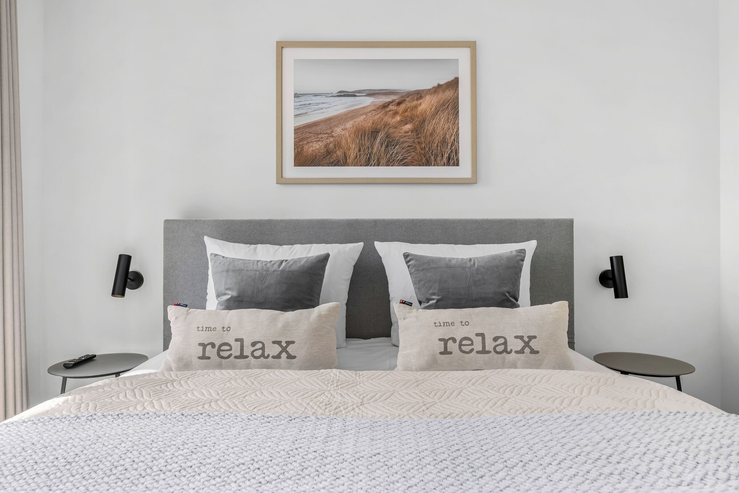 Ein Schlafzimmer mit einem Bett, zwei Nachttischen, einer Wandlampe und einem Bilderrahmen mit einer Strandlandschaft.