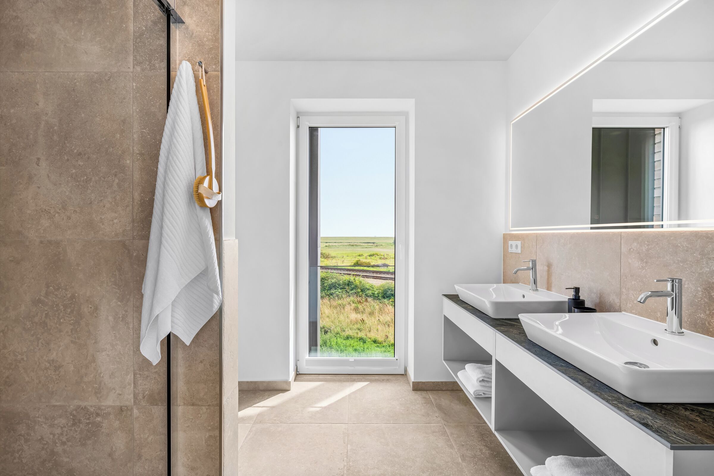A modern bathroom with two sinks, a large mirror, and a view of a green landscape through a door.