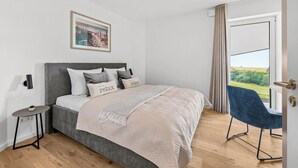 Een slaapkamer met een bed, nachtkastje, stoel, raam met gordijnen en een aan de muur hangend schilderij.