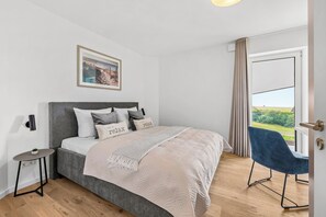 Ein Schlafzimmer mit einem Bett, einem Nachttisch, einem Sessel, einem Fenster mit Vorhängen und einem Wandbild.