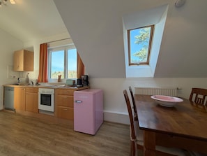 Een keuken met houten kastjes, een roze koelkast, een eettafel met stoelen en een raam met uitzicht op bomen.