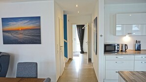 Moderni keittiö, jossa on valkoiset kaapit, mikroaaltouuni ja vedenkeitin. Eteisestä on kulku huoneeseen, josta avautuu näkymä auringonlaskuun.