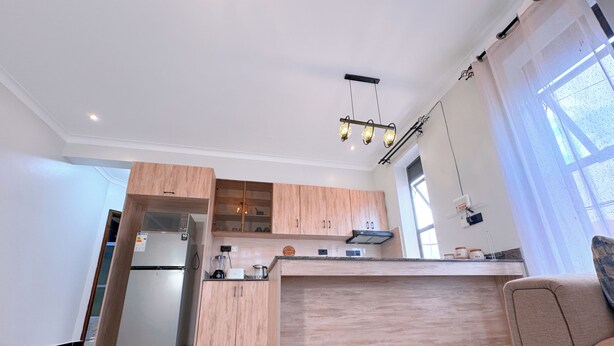 A modern kitchen with wooden cabinets, a built-in oven, a coffee machine, and a dining area with a light-colored sofa. - Constatia Homes Bunga (Kampala)