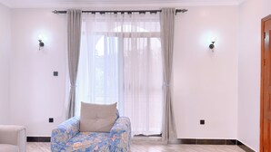 A room with a floral-patterned sofa, a wooden door, and a window with sheer curtains. - Constatia Homes Bunga (Kampala)