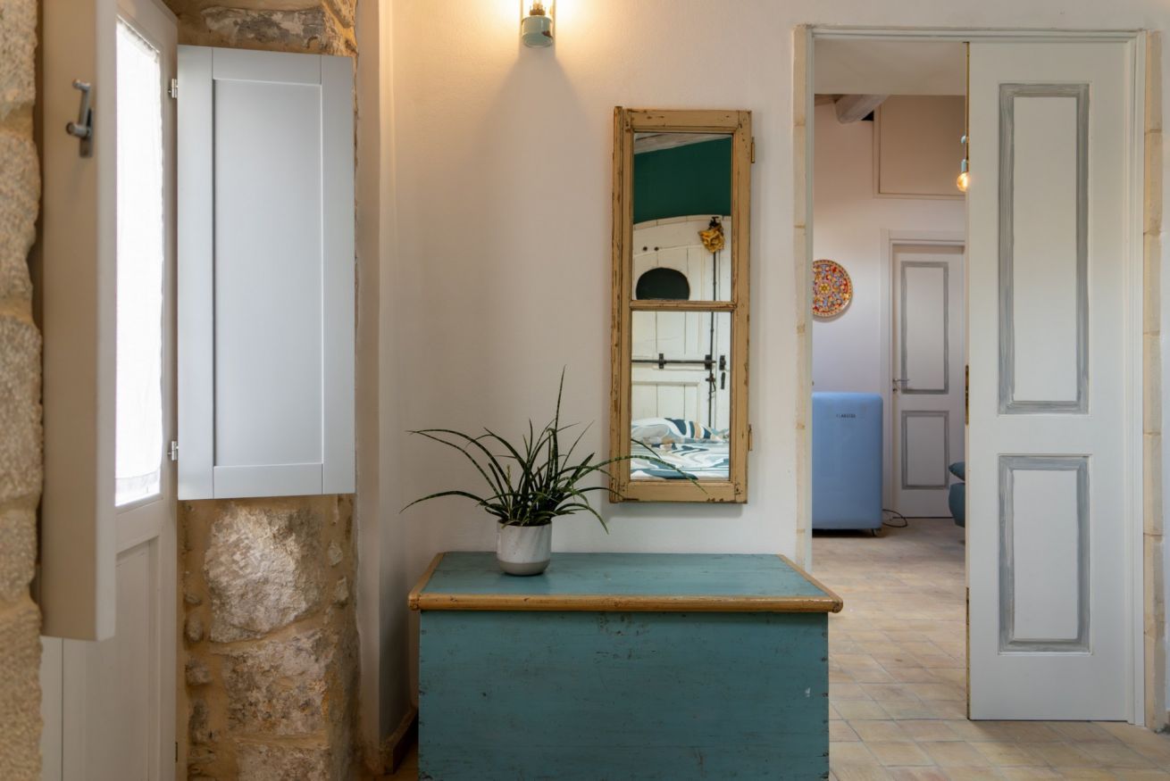 Un couloir avec un mur en pierre, une commode turquoise ornée d’une plante, un miroir et une porte donnant sur une autre pièce.