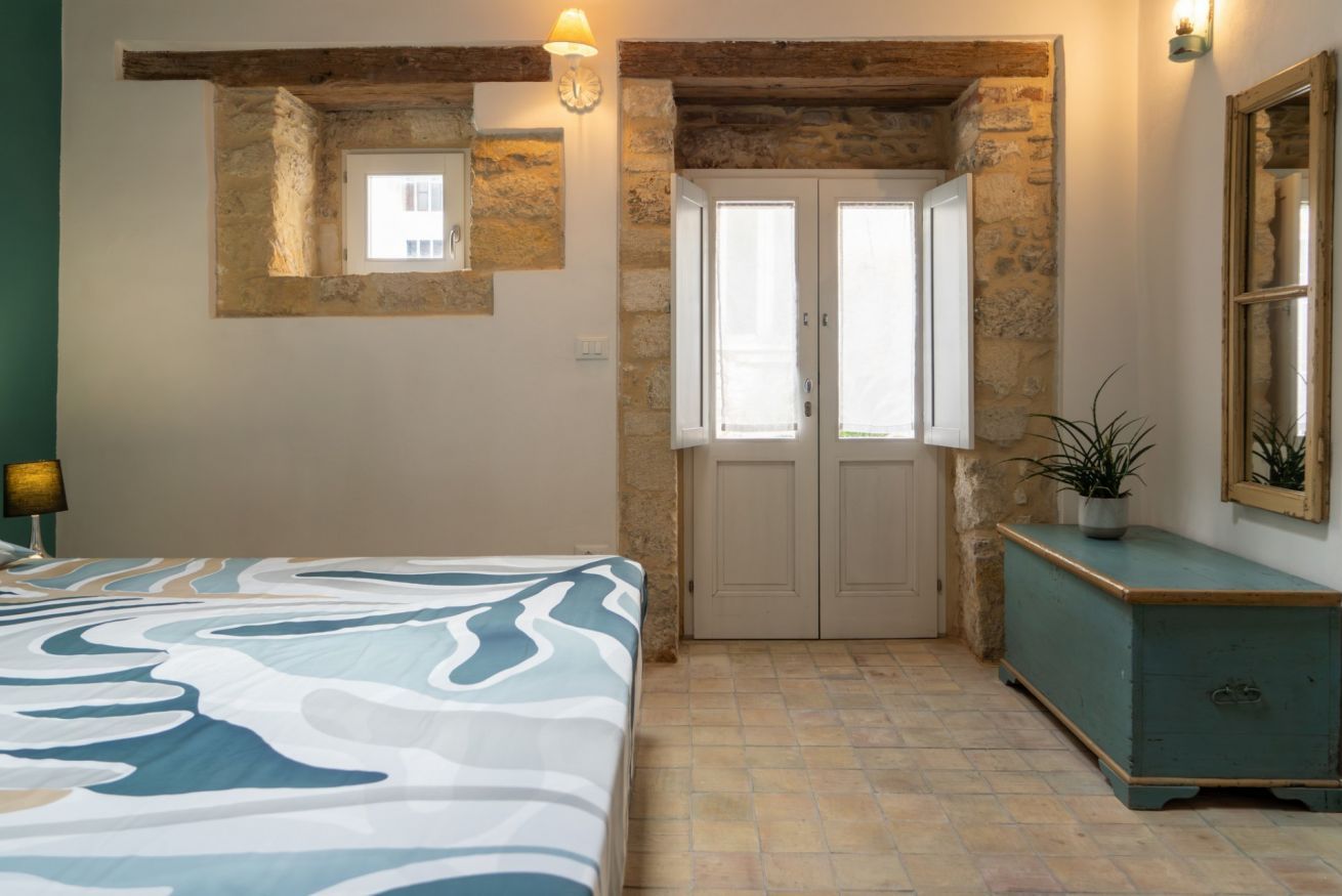 A bedroom with a bed, a stone wall, a window, a door, a bedside table, and a nightstand.