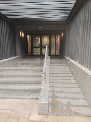 Eine Treppe führt zu einem Gebäudeeingang mit einem Metallgeländer, einem Schild über den Türen und einer gemusterten Decke.