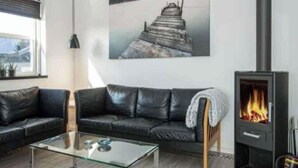 A living room with a glass coffee table, a black leather sofa, a painting of a pier, and a wood-burning stove. - 4 Star Holiday Home in Juelsminde (Juelsminde)