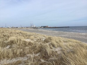 A coastal landscape with a sandy beach, grassy dunes, and industrial structures in the background. - 4 Person Holiday Home in Grenaa (Grenaa)