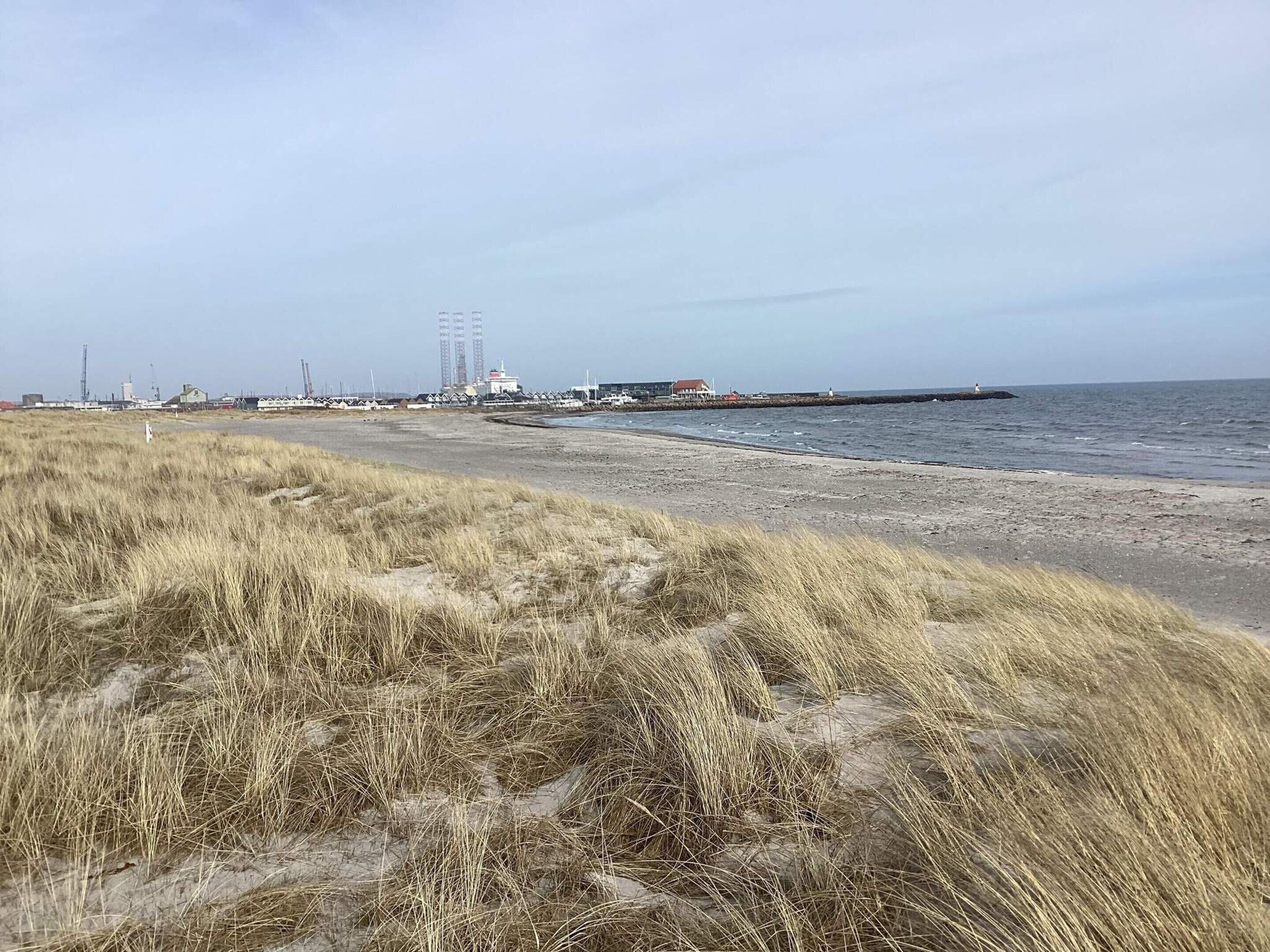 Uma praia com dunas gramadas, um píer que se estende sobre a água e estruturas industriais ao fundo.