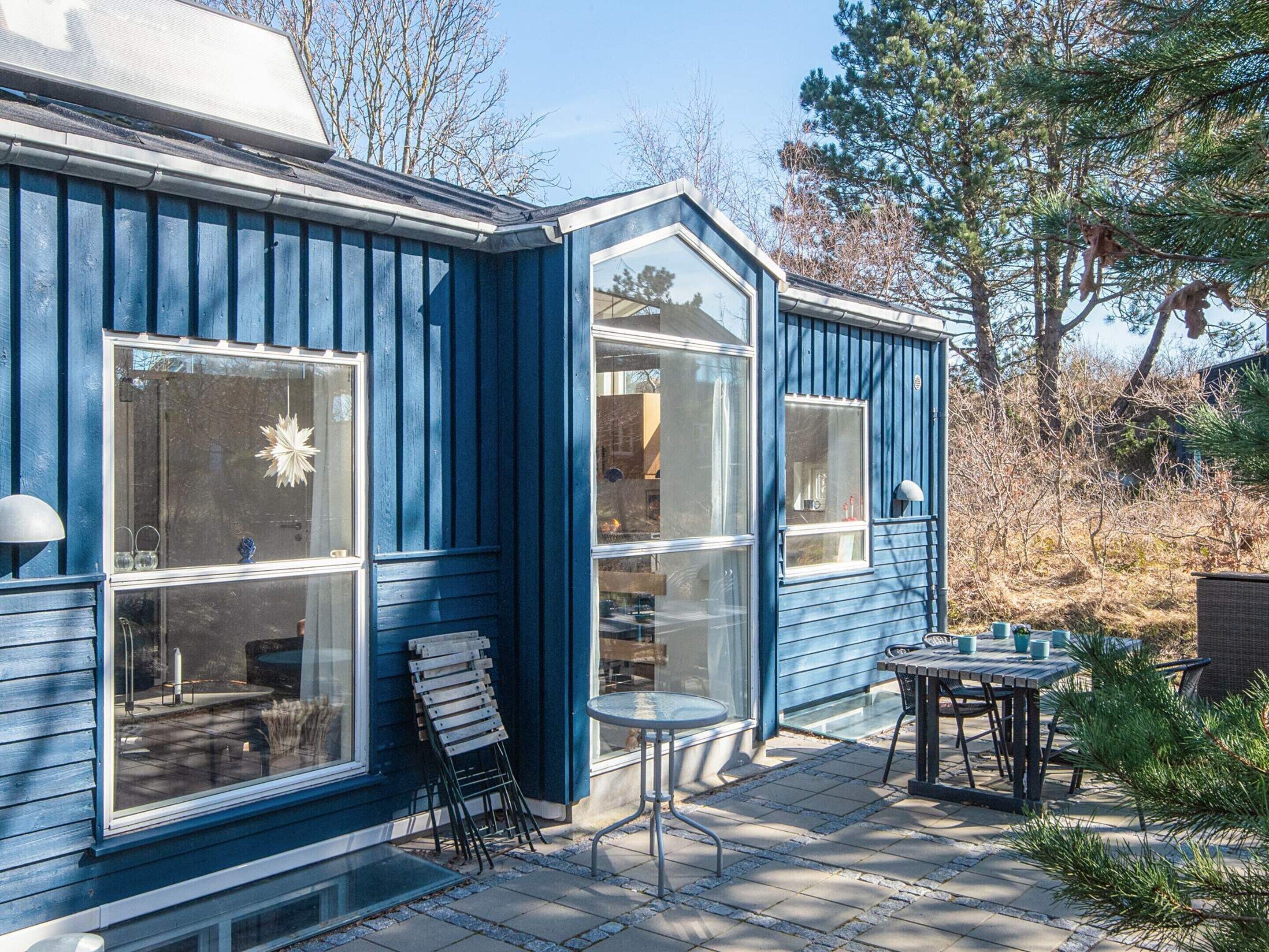 Uma casa azul com janelas amplas, um pátio com mesa e cadeiras, e um pequeno jardim.