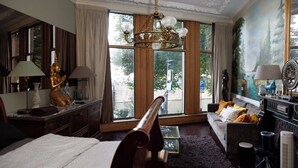 Hotelkamer met een bed, een bank, een bureau, een kroonluchter en grote ramen met uitzicht op bomen.