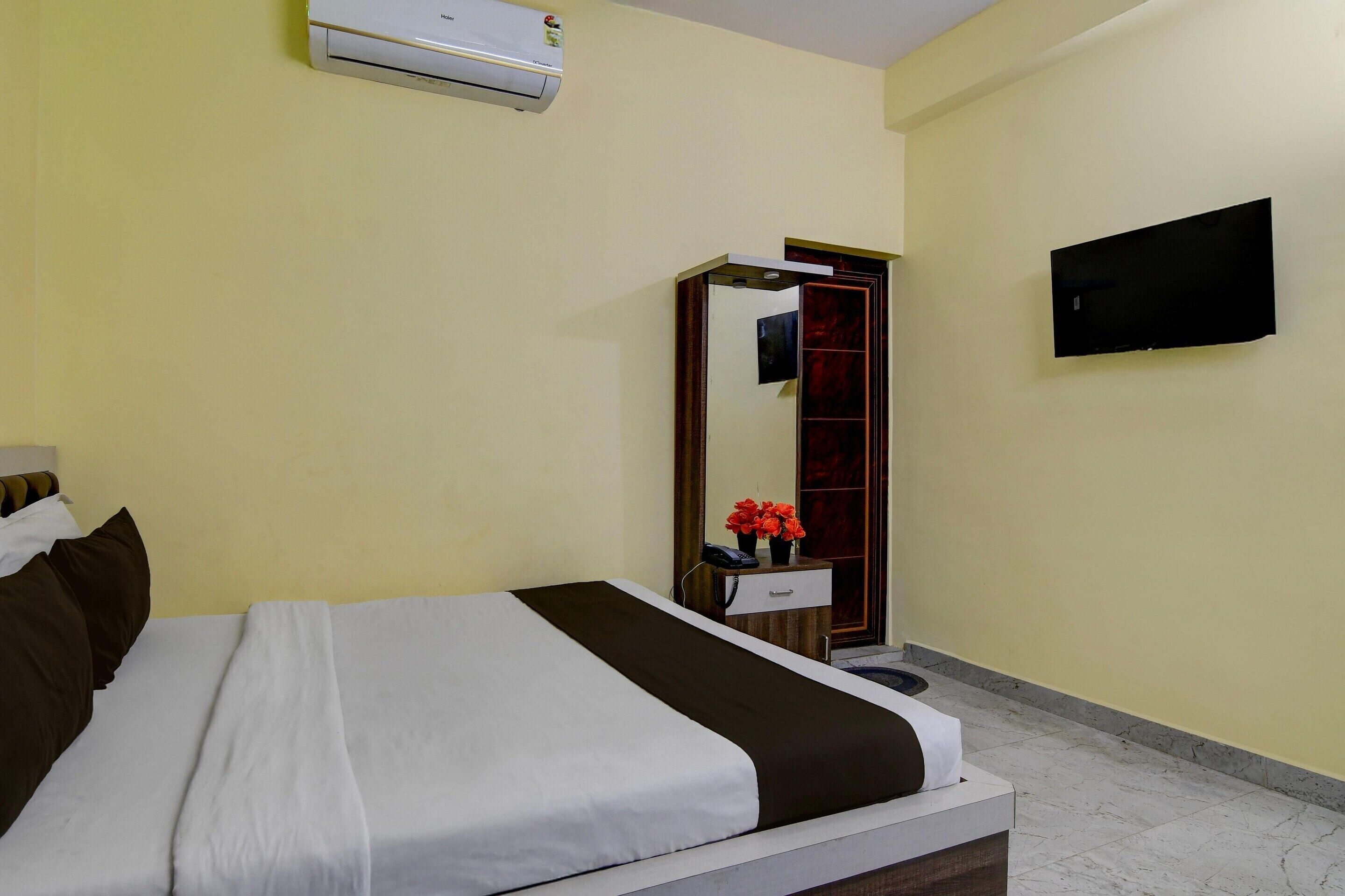A hotel room with a bed, a bedside table, a mirror, an air conditioner, and a TV mounted on the wall.