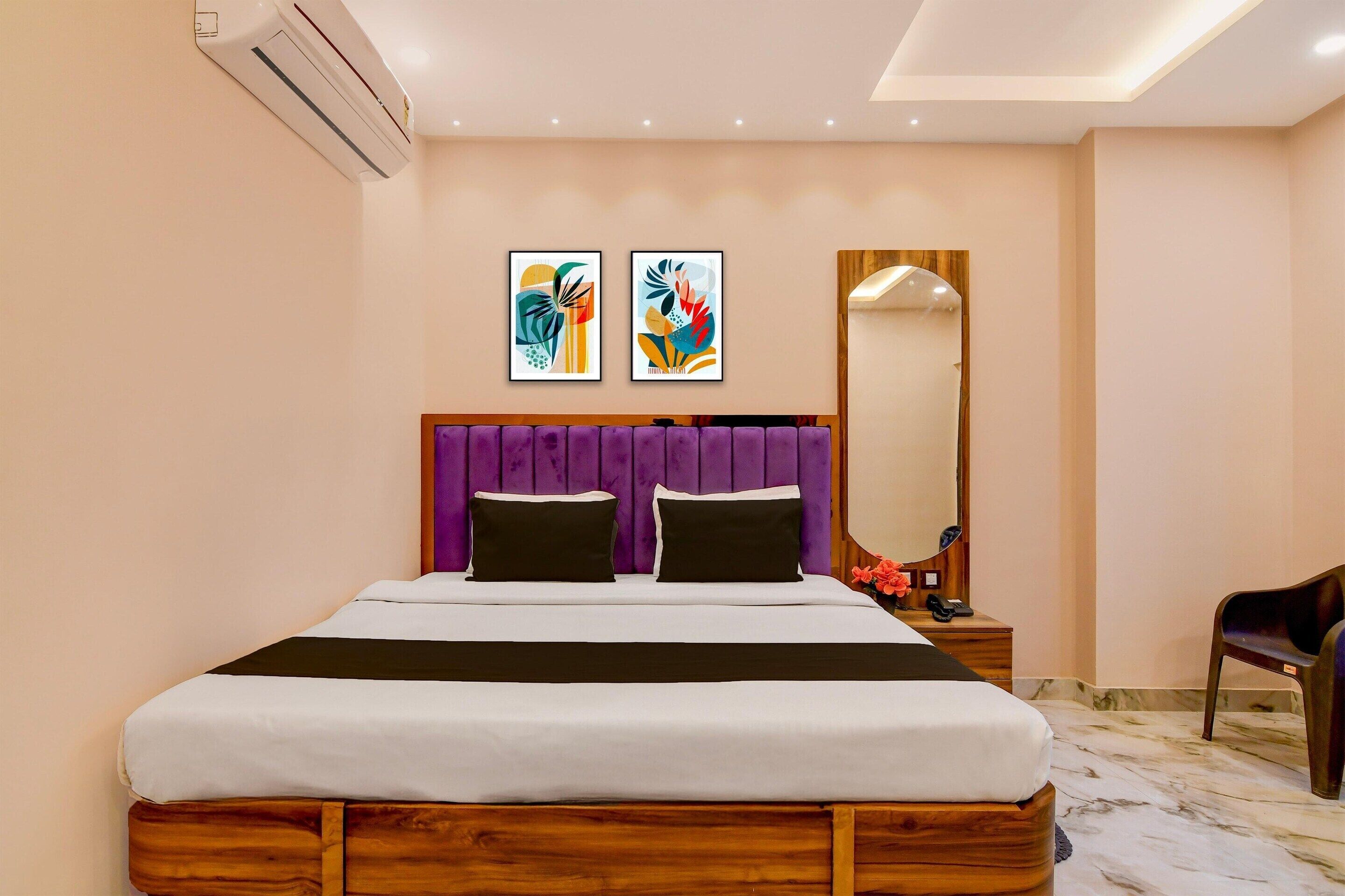 A modern hotel room with a wooden bed frame, a purple headboard, two black pillows, a white bedspread, a wooden nightstand with a phone, and a mirror.