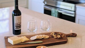 Un piano di lavoro della cucina con una bottiglia di vino, due bicchieri, un tagliere con formaggio e frutta secca, e una candela accesa.