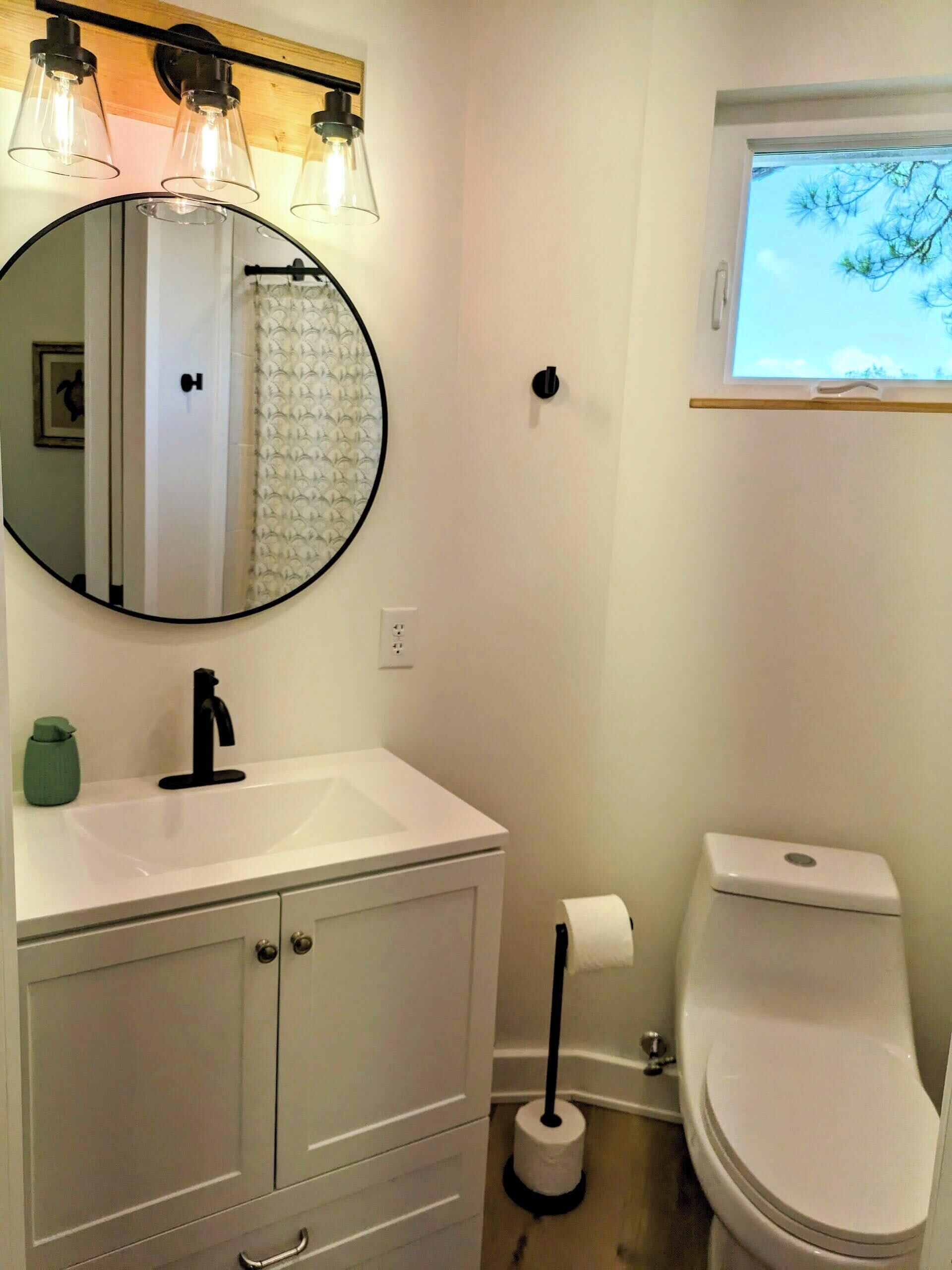 A bathroom with a white sink, a round mirror, a toilet, and a window with a view of trees.