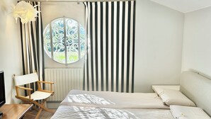 Un dormitorio con cama, una silla, una mesa, un ventanal con cortinas y un radiador.