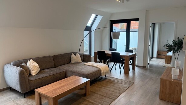 A modern living room with a sectional sofa, a wooden coffee table, a dining area with a table and chairs, and a large window providing natural light. - City Apartment With Terrace (Hasselt)