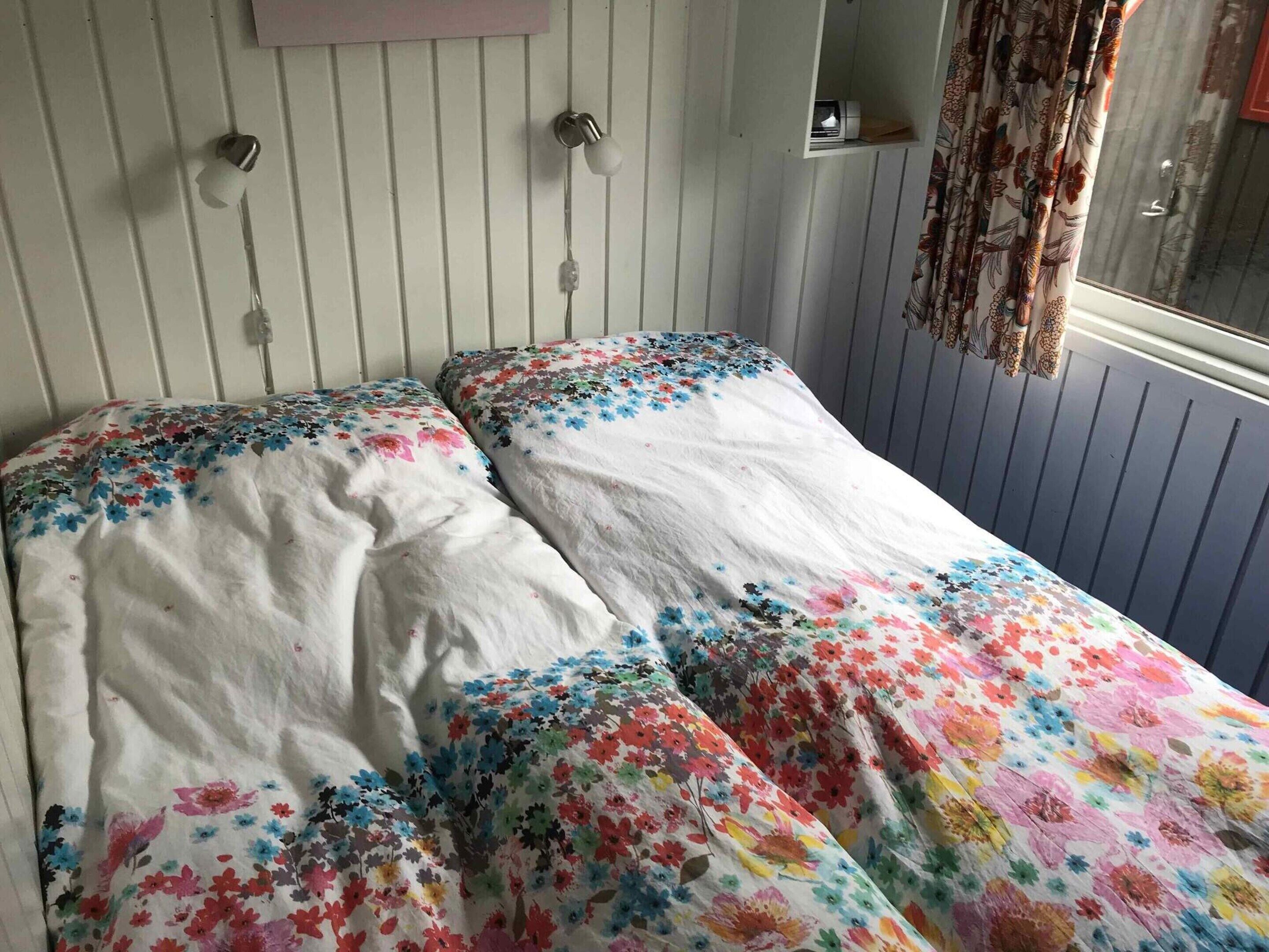 A room with two beds, a window with curtains, a shelf with items, and a wall-mounted light fixture.